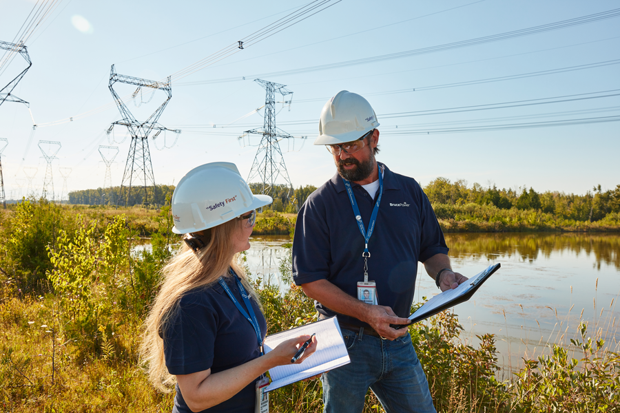 Photograph of a man and woman with hard helmets on going through some documents while outside in front of a lake and powerlines in the background.