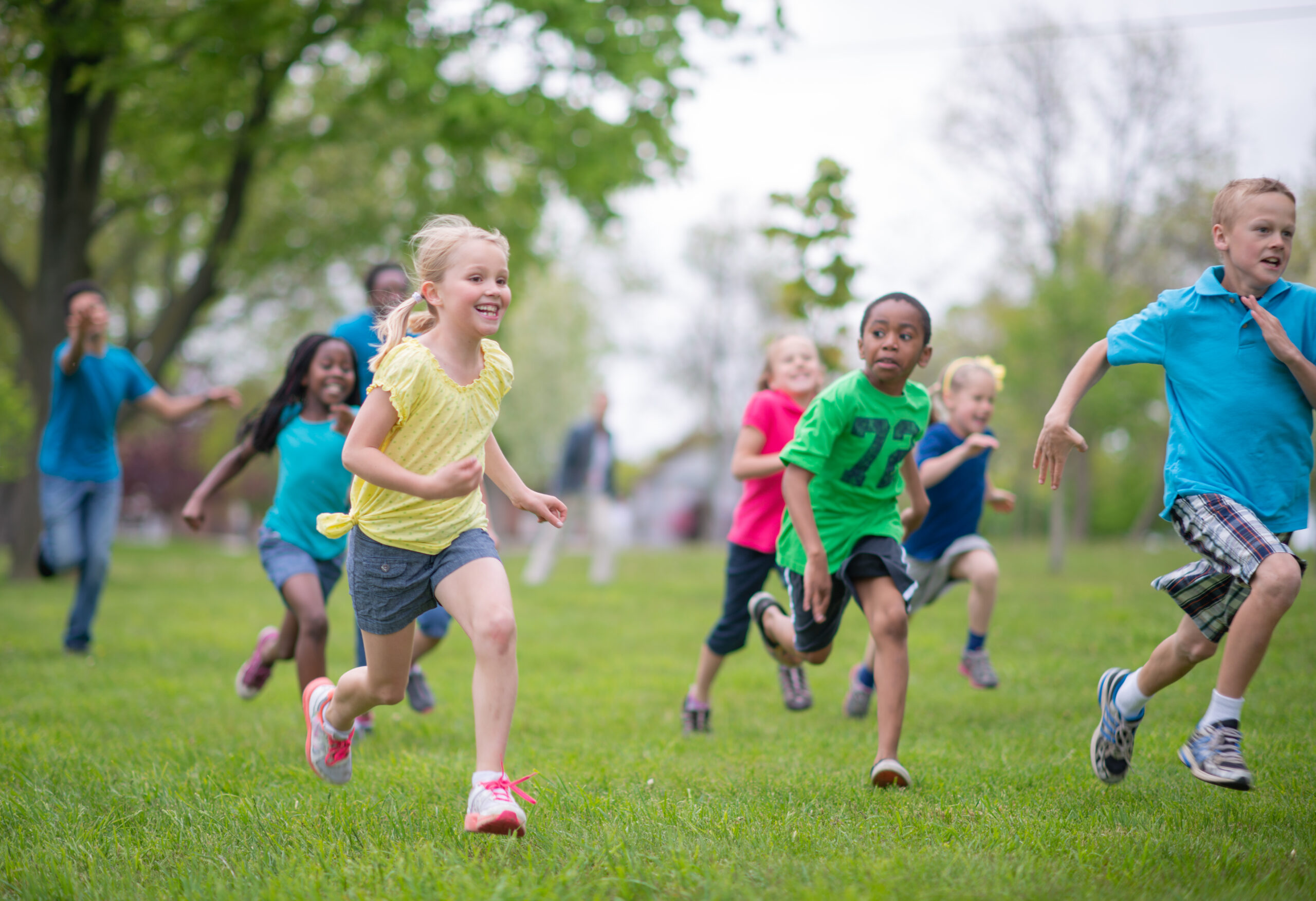 A photograph of a group of children running outdoors at a public park together. They are dressed casually on a summer's day.